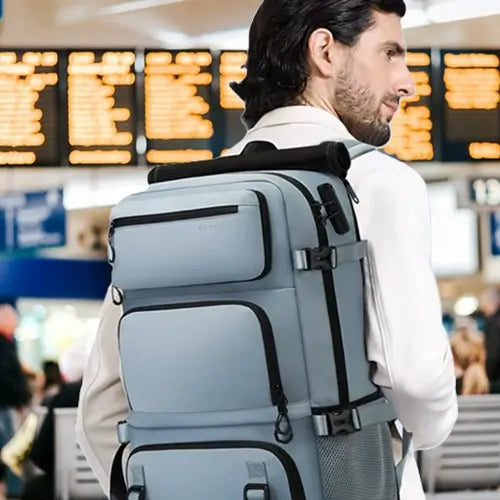 Homme barbu de profil, portant un grand sac à dos de voyage gris clair avec multiples compartiments et bretelles renforcées, debout dans un hall d’aéroport animé, face à un panneau lumineux affichant les départs.