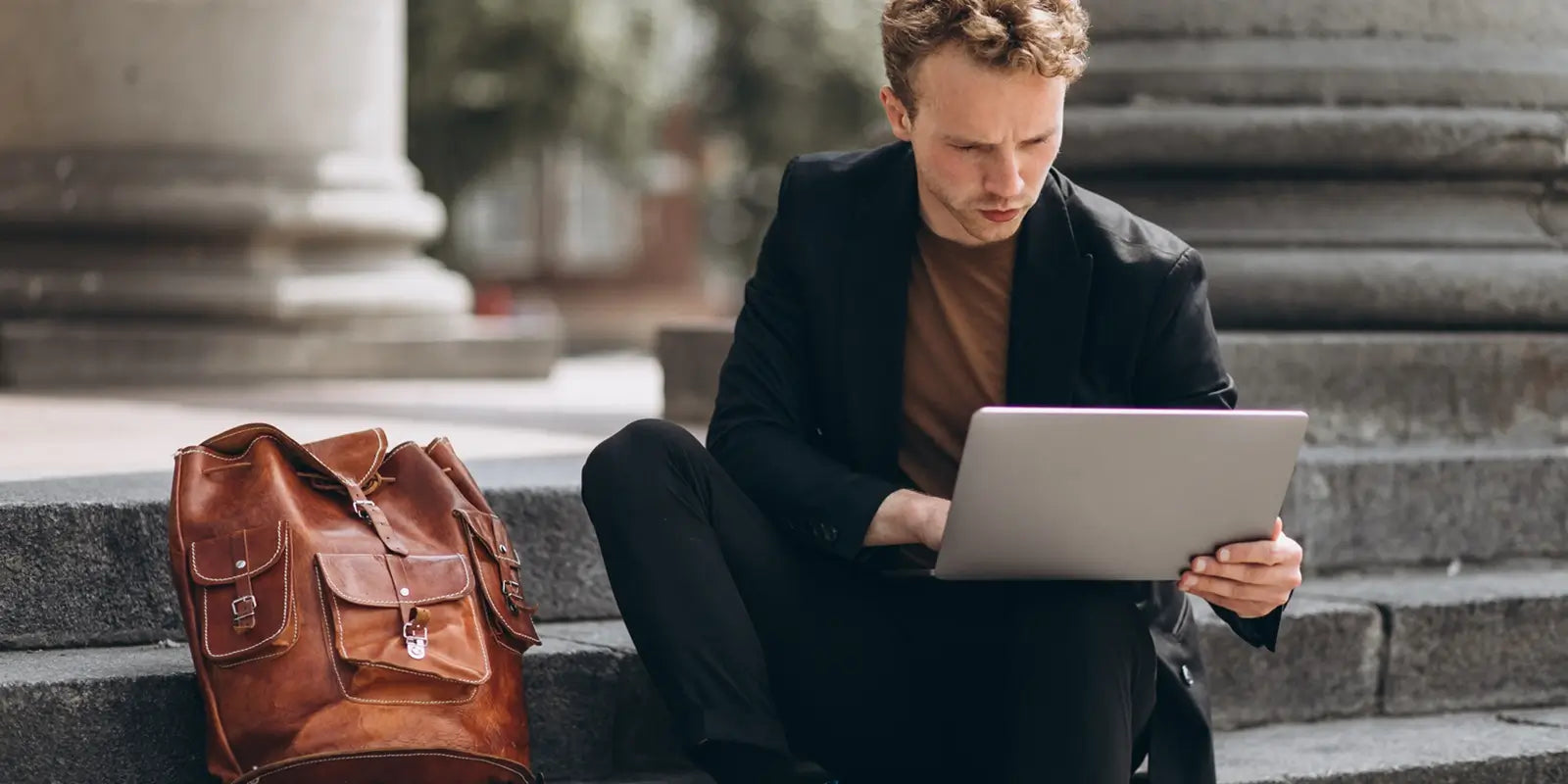 Jeune homme assis sur les marches d’un bâtiment en pierre, concentré sur son ordinateur portable, avec un sac à dos homme en cuir marron posé à côté de lui, illustrant un mode de vie nomade et connecté.