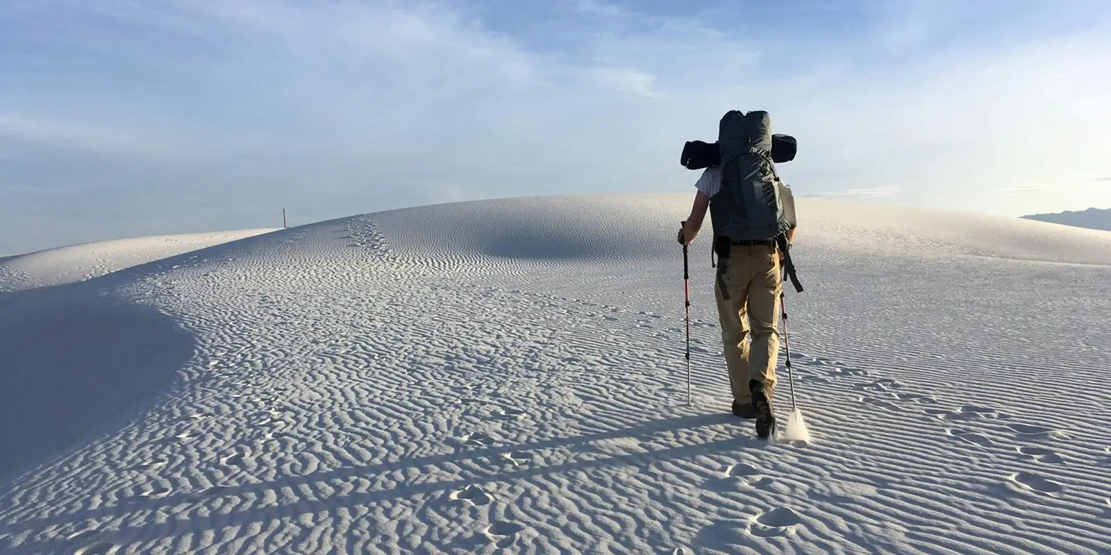 Sac à dos Sport - Randonneur équipé marchant sur des dunes blanches au lever du soleil, sac de trekking volumineux et bâtons de marche visibles