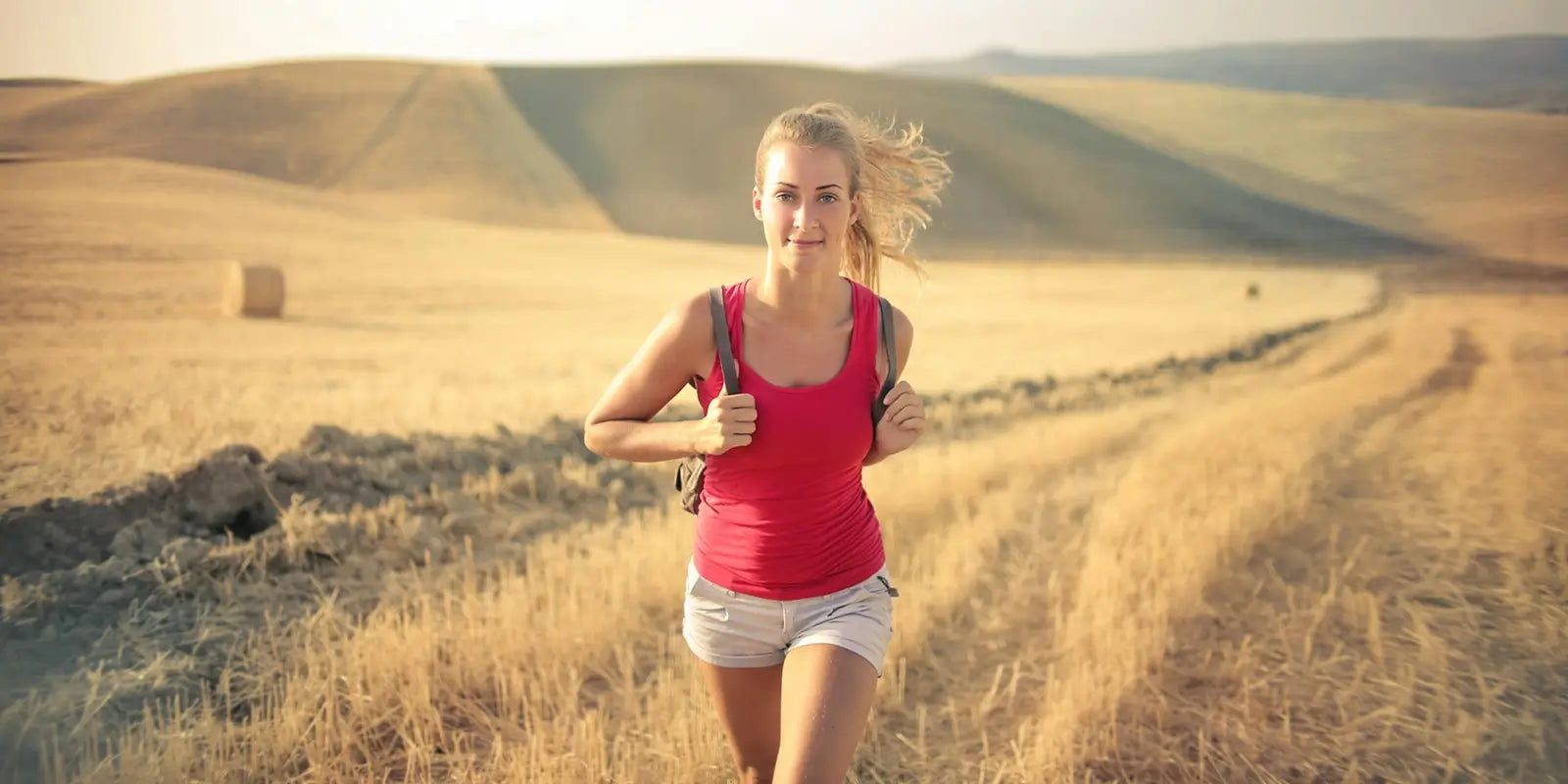 Sac à dos Sport - Jeune femme courant dans un champ doré avec un petit sac à dos de randonnée, paysage vallonné en arrière-plan.