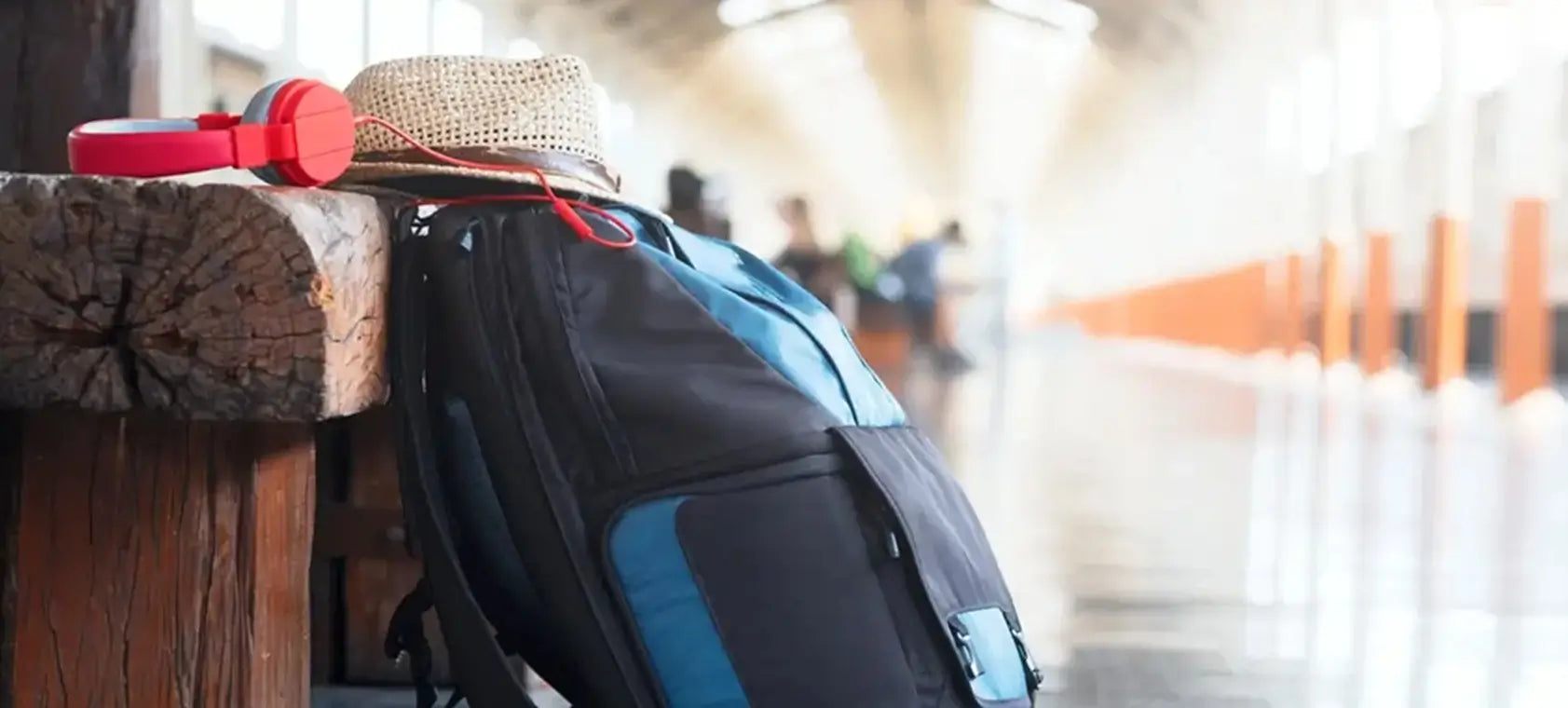 Sac à dos de voyage noir et bleu appuyé sur un banc en bois dans une gare, avec un chapeau de paille et un casque audio rouge posés dessus
