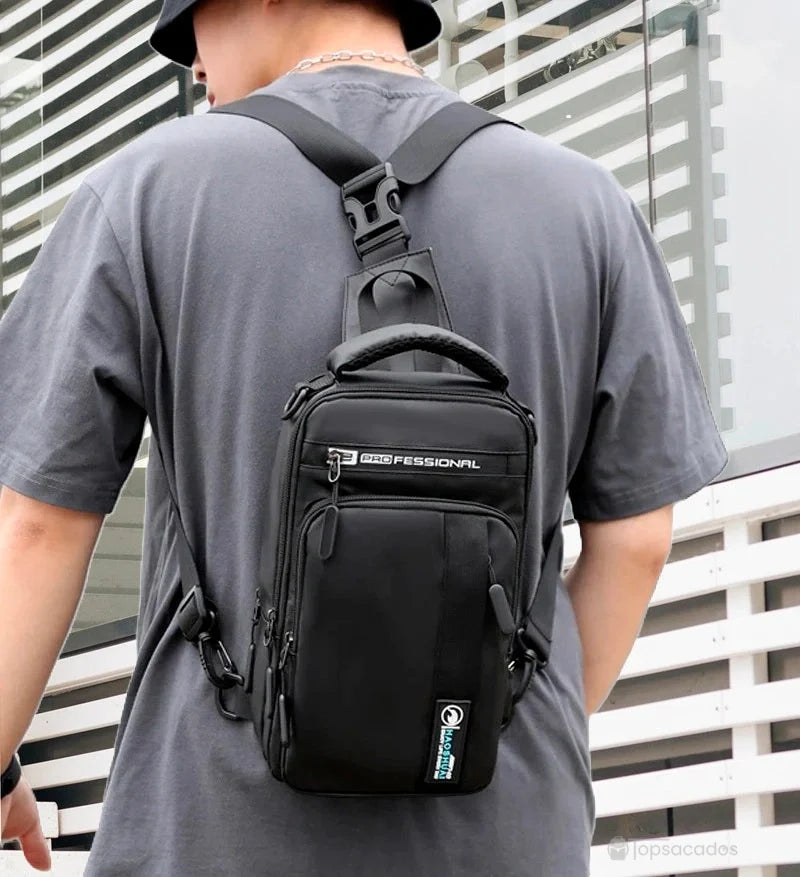 Petit sac à dos homme noir porté avec sangle croisée sur le dos, devant une façade vitrée moderne, style casual.