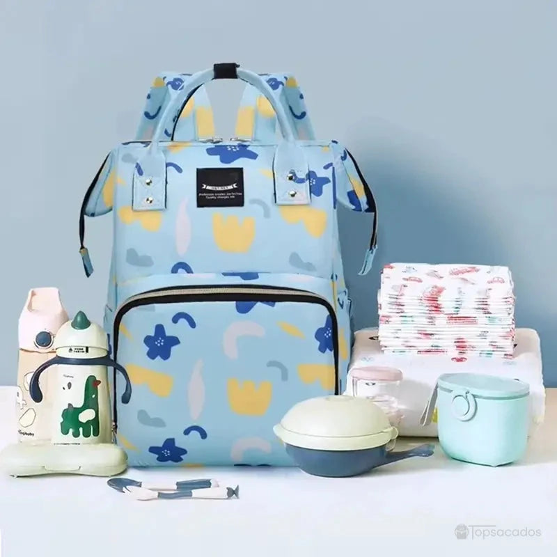 Sac à dos à langer bleu clair entouré d’accessoires de puériculture comme des couches, des boîtes repas et des biberons, sur fond bleu clair.