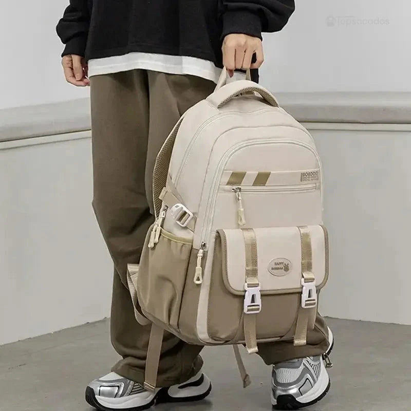 Sac à dos ado beige et kaki porté à la main par un adolescent habillé en vêtements décontractés, en position debout