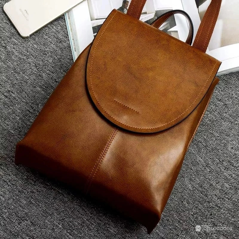 Sac à dos cuir femme marron posé au sol sur tapis gris avec magazine et téléphone à proximité, ambiance élégante et cosy.