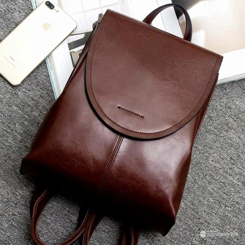 Sac à dos cuir femme marron foncé posé sur moquette grise, ambiance moderne avec téléphone et magazine en arrière-plan.