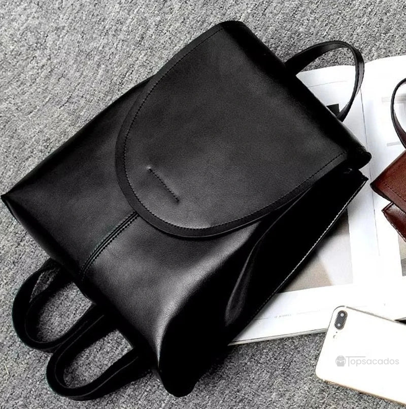 Sac à dos femme en cuir noir allongé sur une surface grise à côté d’un smartphone, illustrant sa texture lisse et sa praticité urbaine.