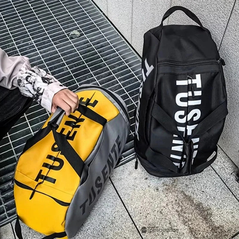Sac à dos foot noir posé debout à côté d’un sac jaune et gris tenu à la main, sur sol en béton extérieur.