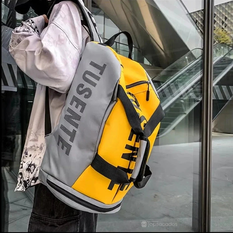 Sac à dos foot jaune et gris porté sur le dos en extérieur urbain, devant une façade vitrée contemporaine.