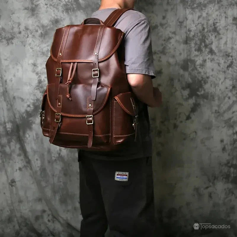 Sac à dos homme cuir brun foncé porté sur l'épaule en intérieur contre un mur texturé gris