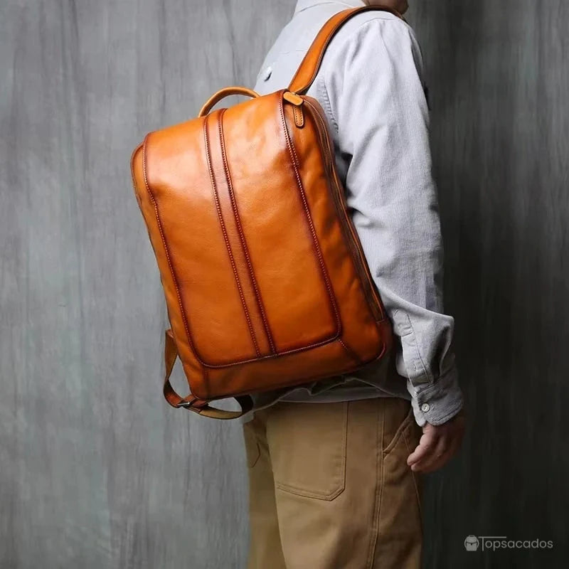 Sac à dos homme cuir marron porté à l'épaule par un homme habillé en chemise grise et pantalon beige, ambiance intérieure sobre.