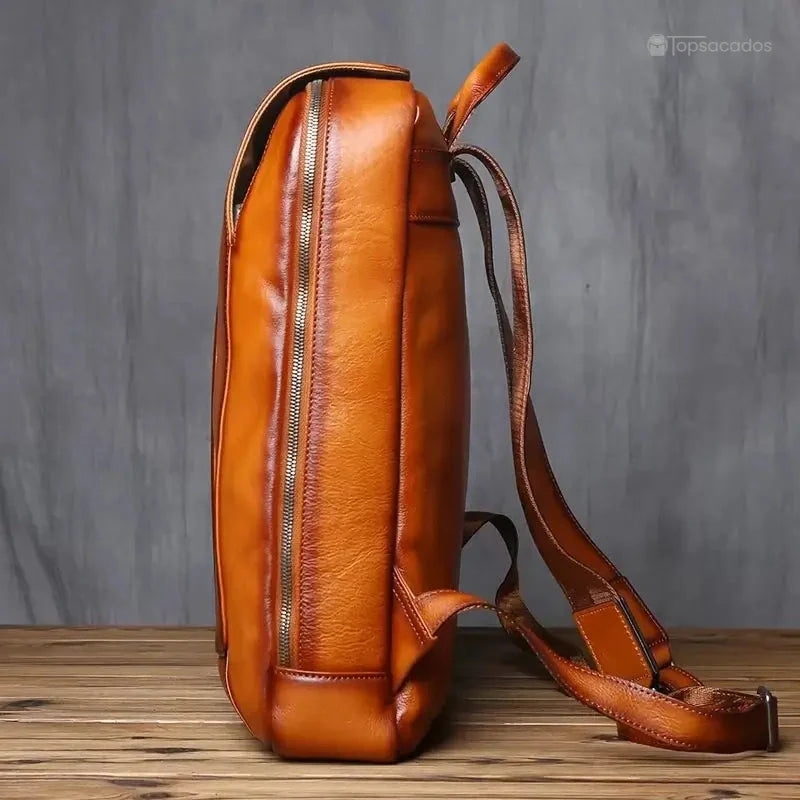Sac à dos homme cuir marron vu de profil, mettant en valeur l'épaisseur du cuir et la qualité de la couture sur fond bois et mur gris.