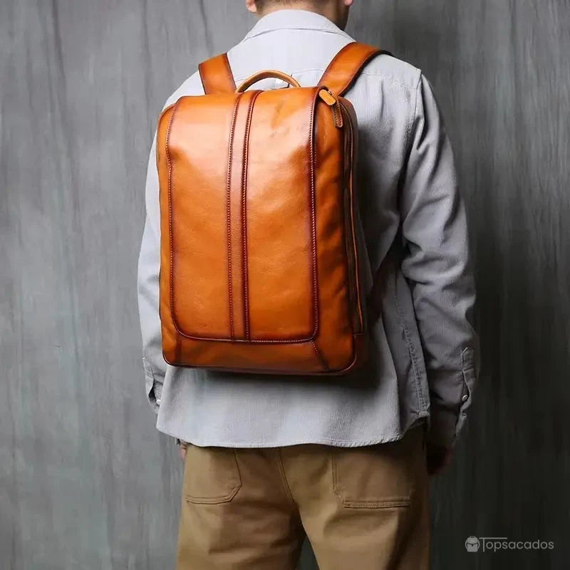 Sac à dos homme cuir marron porté dans le dos par un homme, vue arrière complète dans un environnement intérieur au mur gris clair.