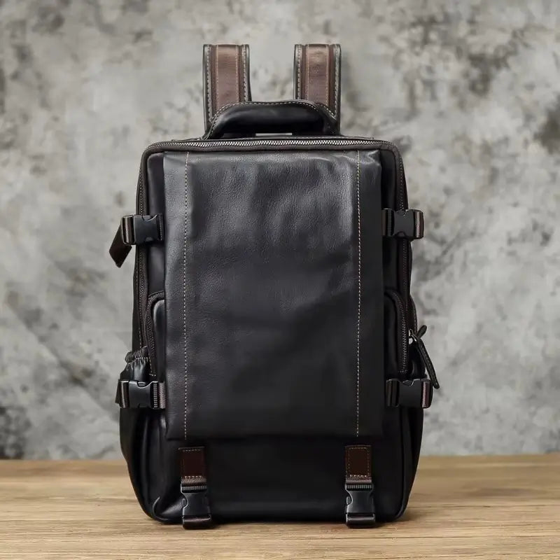 Sac à dos homme cuir noir vu de face avec bretelles visibles, posé sur une table en bois devant un mur bétonné.