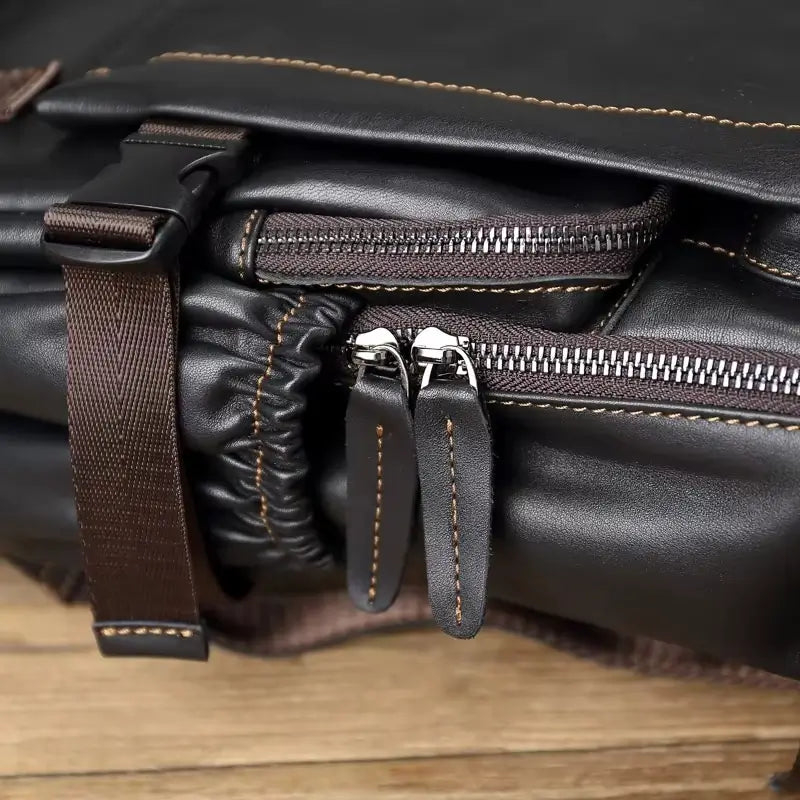 Sac à dos homme cuir noir gros plan sur les fermetures éclair robustes et les sangles marron sur fond de bois brut.