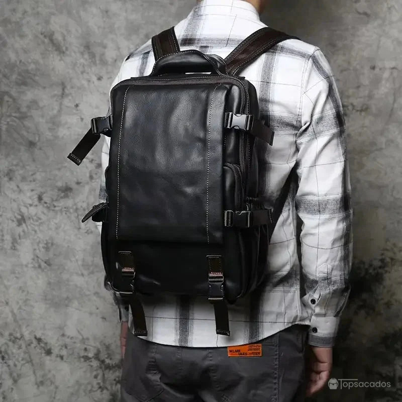 Sac à dos homme cuir noir porté sur le dos d'un homme en chemise à carreaux blancs, fond béton texturé.