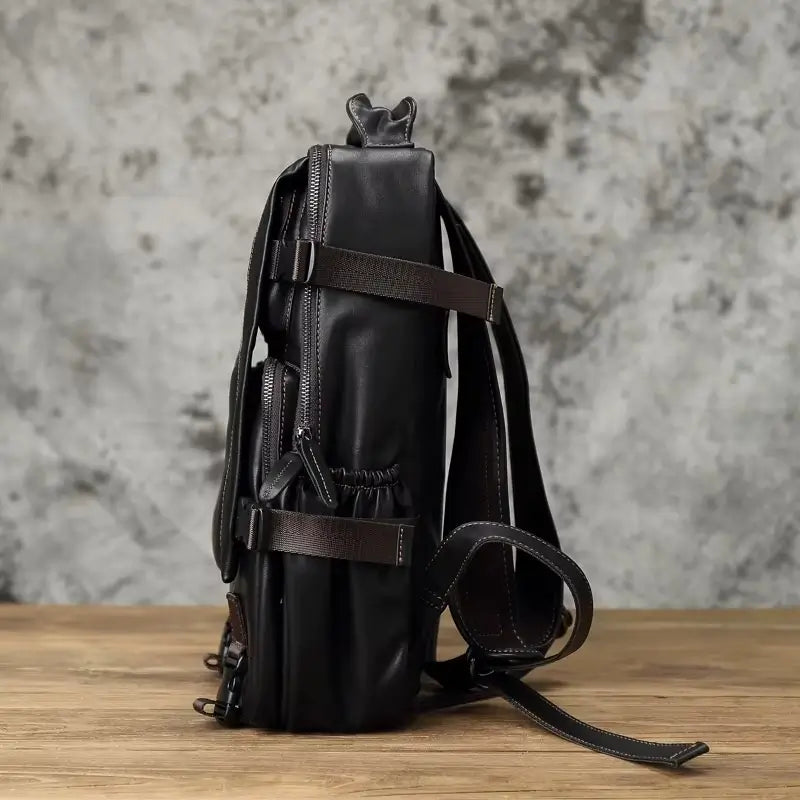 Sac à dos homme cuir noir vue de profil montrant les poches latérales élastiques et les sangles réglables, sur table en bois.