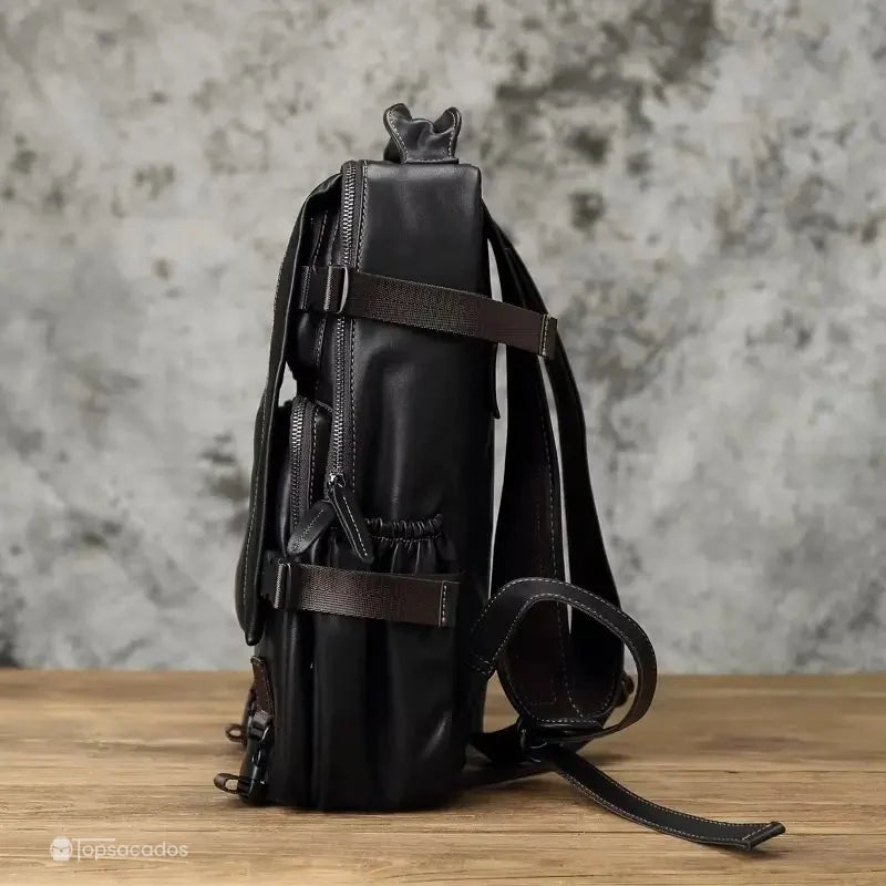 Sac à dos homme cuir noir vue de profil montrant les poches latérales élastiques et les sangles réglables, sur table en bois.