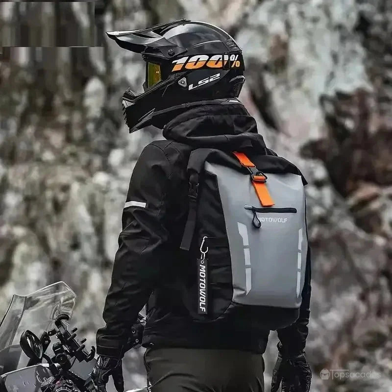 Sac à dos motard porté par un pilote équipé d’un casque cross noir, à l’arrêt devant un paysage de montagne rocheuse.