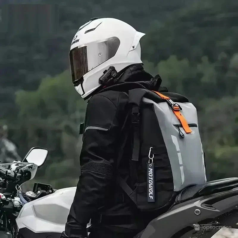 Sac à dos motard porté par un motard en tenue noire et casque blanc, posé sur une moto blanche, en pleine nature.