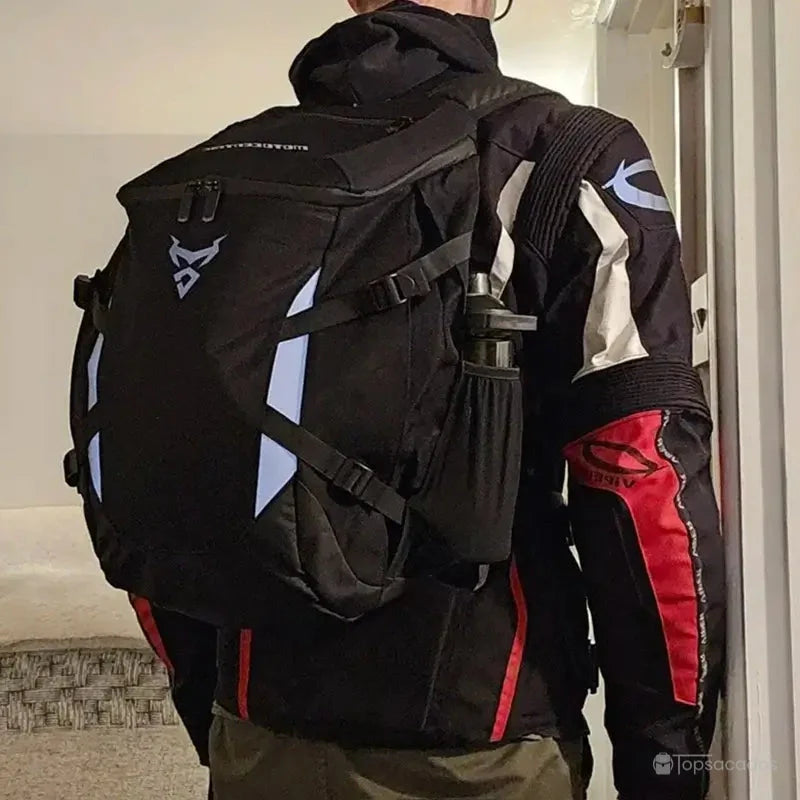 Sac a dos moto étanche porté par un motard debout en tenue complète, photographié en intérieur, avec poches latérales visibles et bouteille fixée.