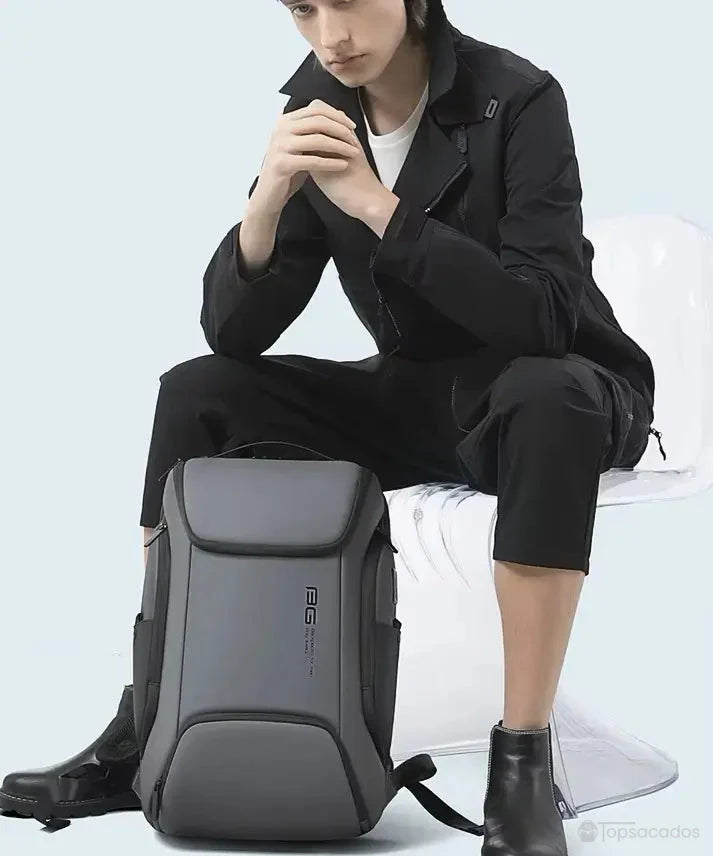 Sac a dos ordinateur portable multipoches gris foncé posé entre les pieds d’un homme assis en tenue noire moderne, illustrant son usage professionnel ou étudiant.