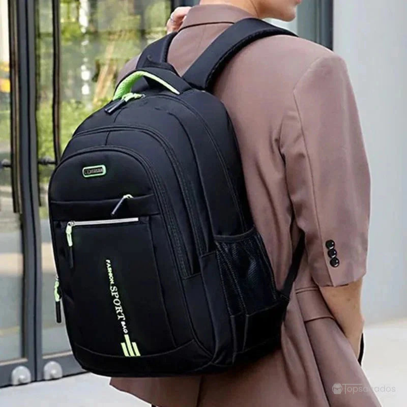 Sac a dos pour cours noir avec finitions vertes porté par une personne en veste beige devant un bâtiment vitré.