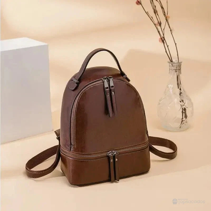 Sac à dos pour femme marron foncé posé sur une surface beige à côté d’un vase en verre transparent et d’un décor minimaliste.