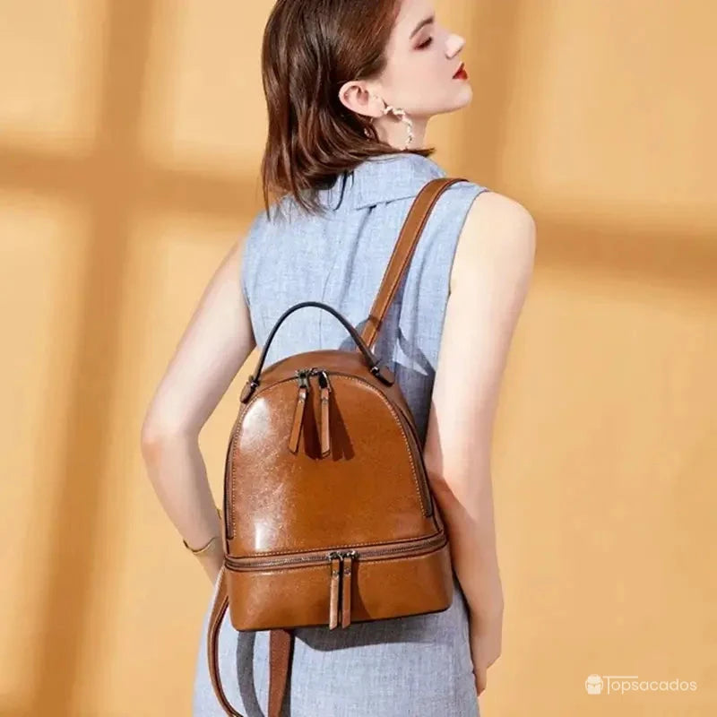 Sac à dos pour femme en cuir marron porté par une femme en robe bleu clair, en intérieur lumineux avec mur orangé.