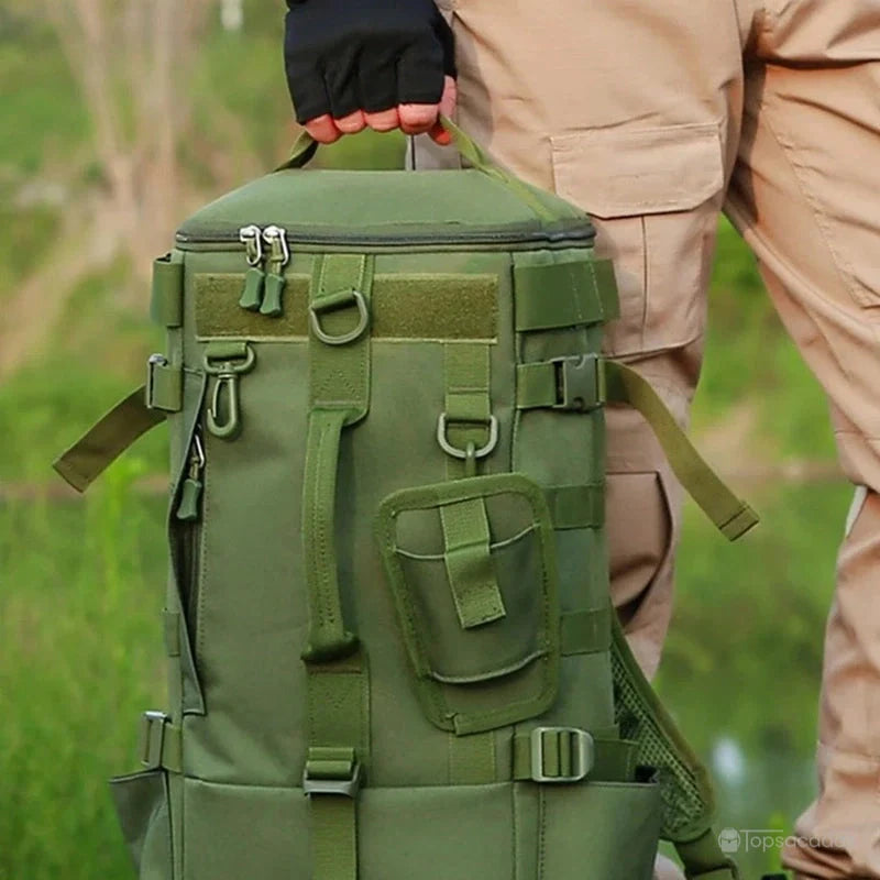 Sac à dos pour la pêche tenu à la main, mis en situation au bord de l’eau, avec vêtements de terrain et végétation en arrière-plan.
