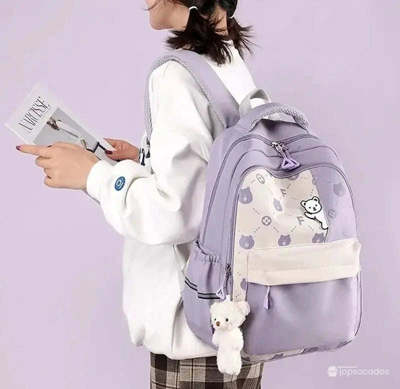 Sac à dos primaire fille violet porté par une élève lisant un magazine sur fond violet clair.