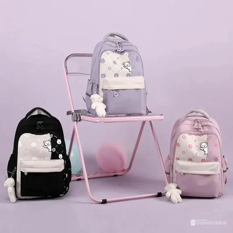 Sac à dos primaire fille en trois couleurs différentes posé sur chaise rose, fond uni violet.