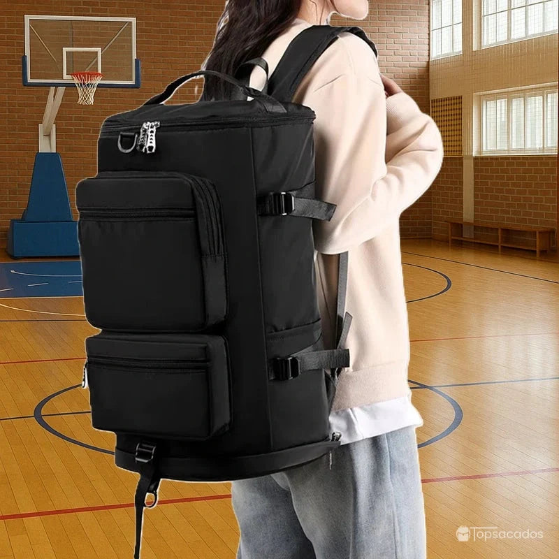 Sac à dos sport grand volume porté par une femme dans un gymnase avec parquet et panier de basket, compartiments multiples visibles.