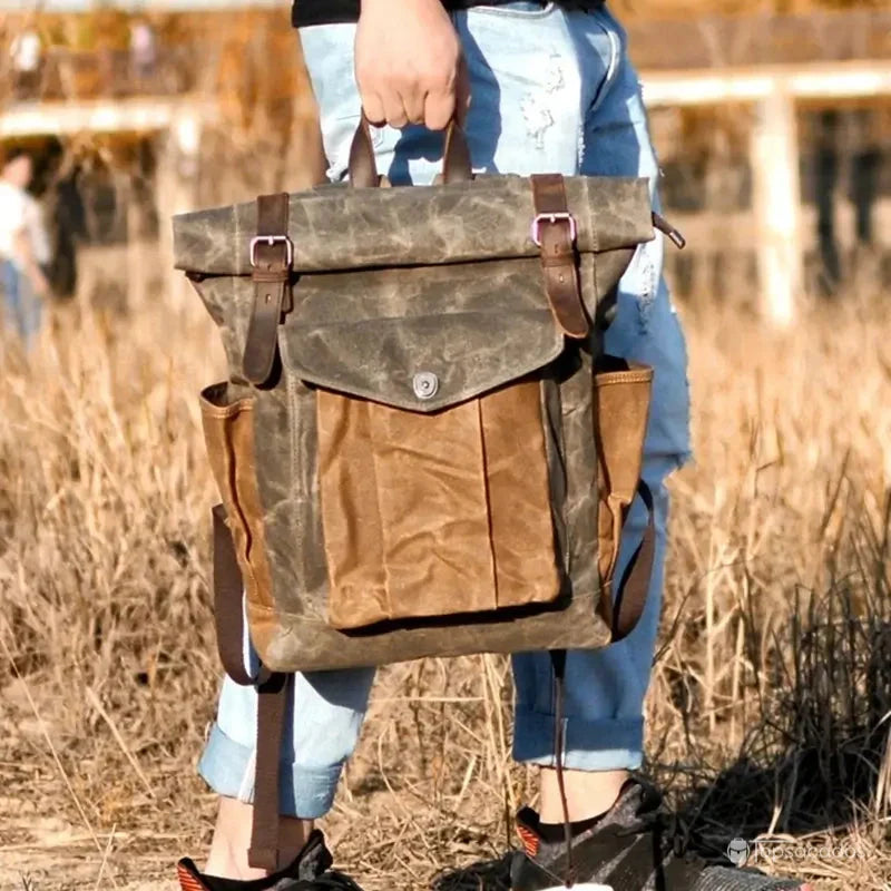 Sac à dos toile homme vintage tenu à la main par un utilisateur en jean dans un champ sec, en pleine nature.