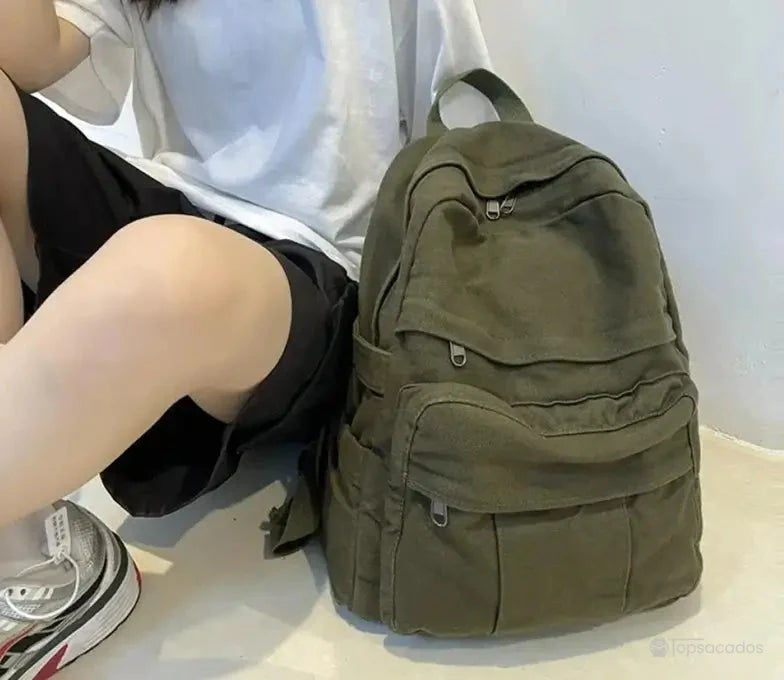 Sac à dos toile vert kaki posé au sol à côté d'une personne assise, ambiance urbaine décontractée