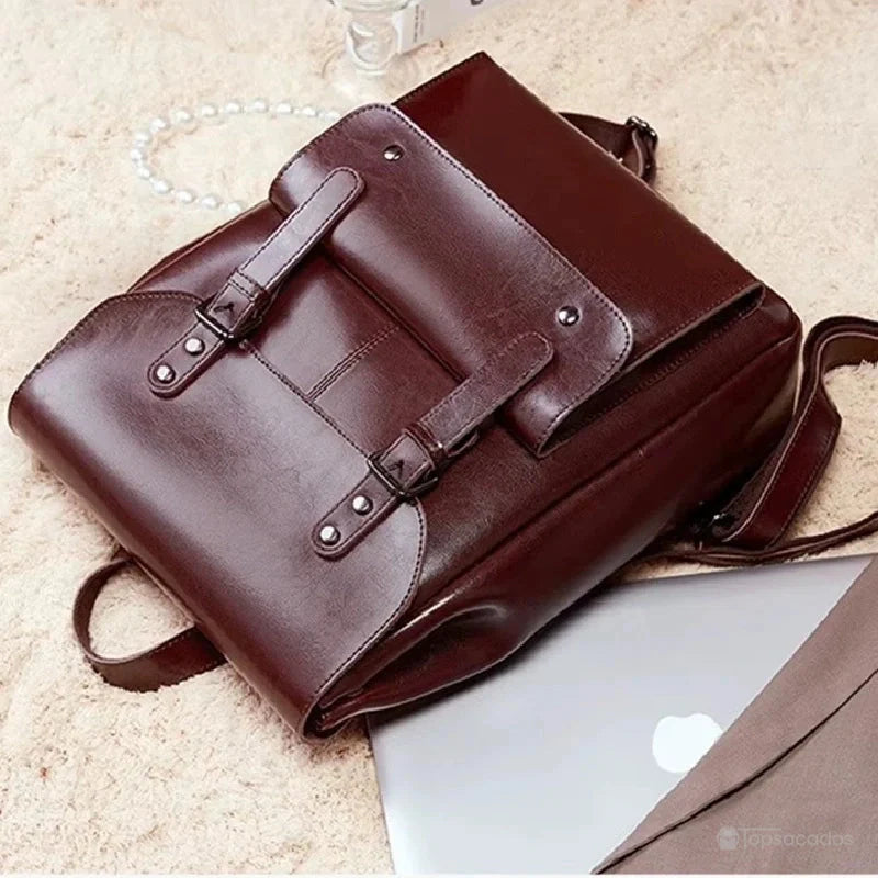 Sac à dos vintage pour femme en cuir marron foncé allongé sur un tapis beige à côté d’un ordinateur portable, suggérant un usage professionnel ou universitaire.