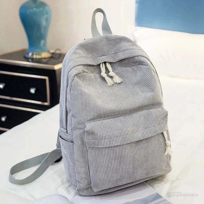 sac à dos college en velours cotelé gris clair avec poches latérales et cordons déco, posé sur un lit