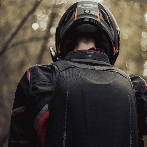 Motard de dos portant un sac à dos moto noir aérodynamique et un casque intégral dans un environnement boisé