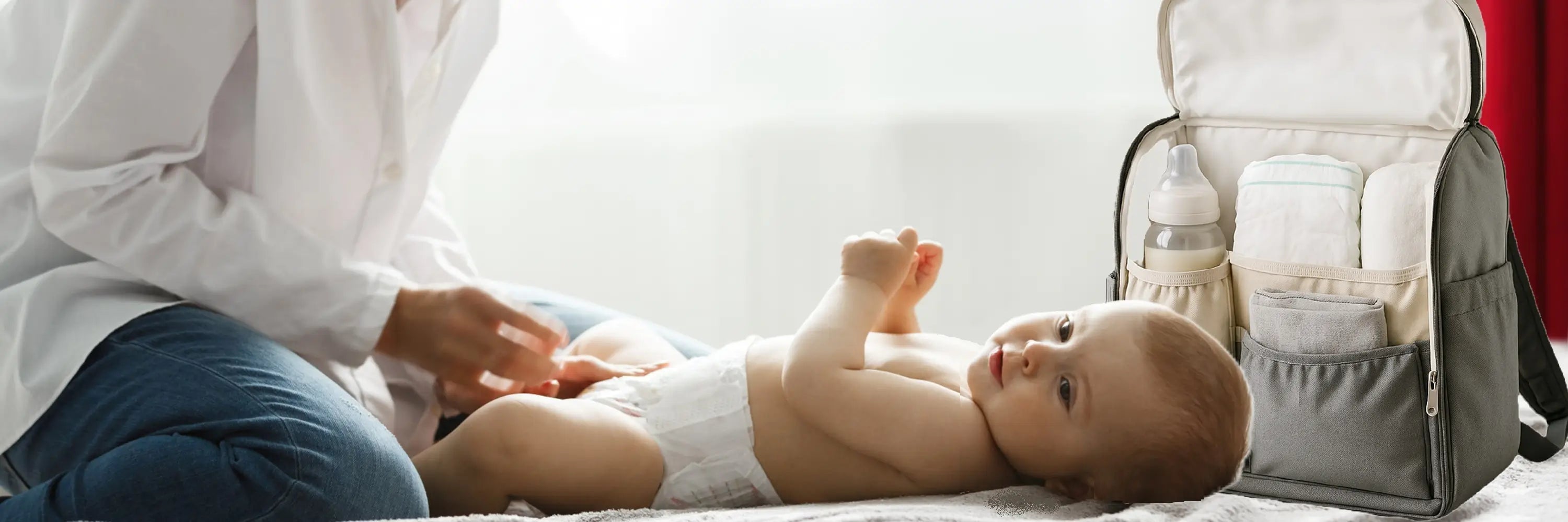 Sac à dos à langer ouvert posé sur un lit à côté d’un bébé allongé en couche, en train d’être changé par un parent. Le sac contient des couches, un biberon, une serviette et d'autres accessoires pour bébé, illustrant un moment de soin pratique et organisé à la maison.