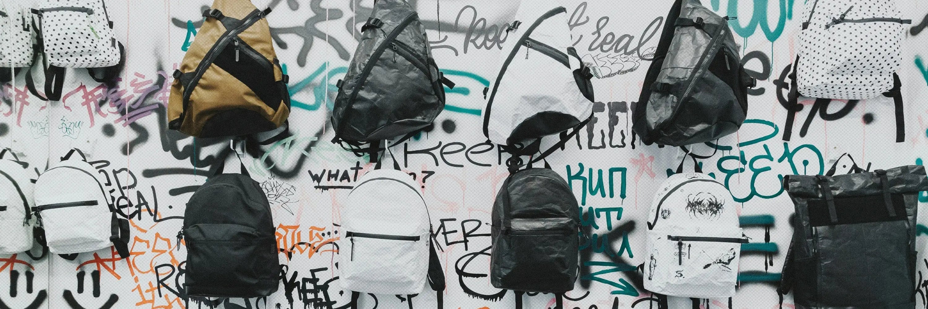 Collection de sacs à dos ecole modernes accrochés sur un mur graffiti, avec différents coloris et designs adaptés au quotidien scolaire et urbain.