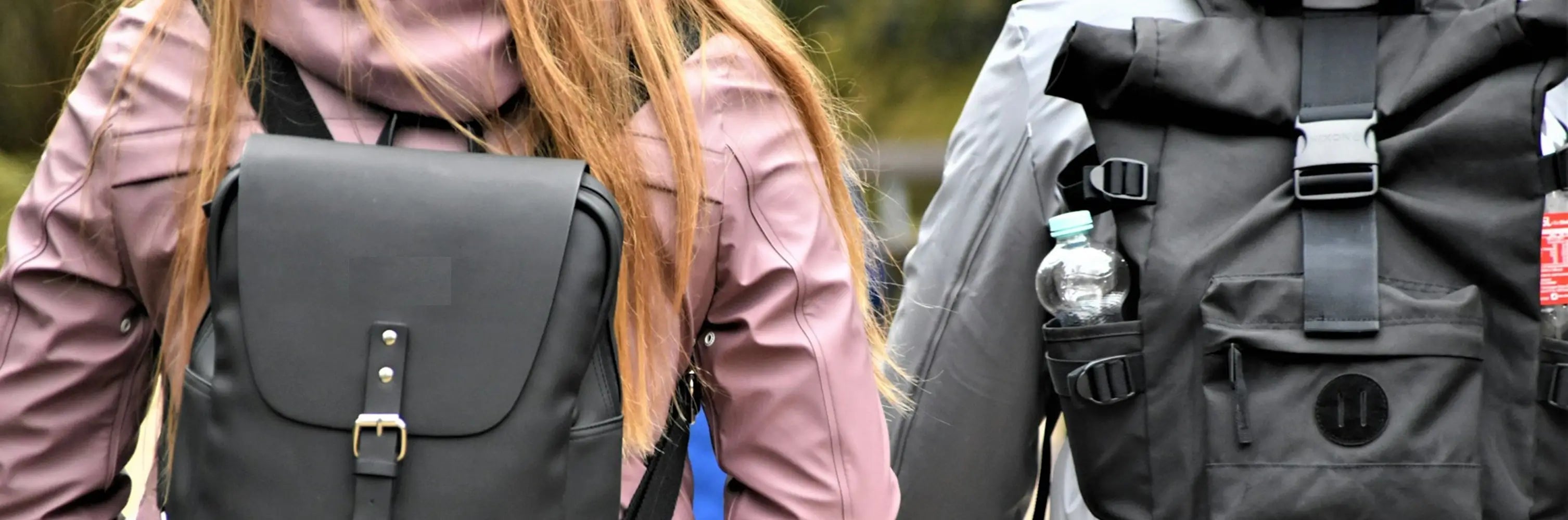 Deux enfants vus de dos portant un sac à dos enfant noir en ville, l’un avec un sac à rabat classique et l’autre avec un sac à dos roll-top, bouteille d’eau visible sur le côté, marchant côte à côte dans un environnement urbain en extérieur.