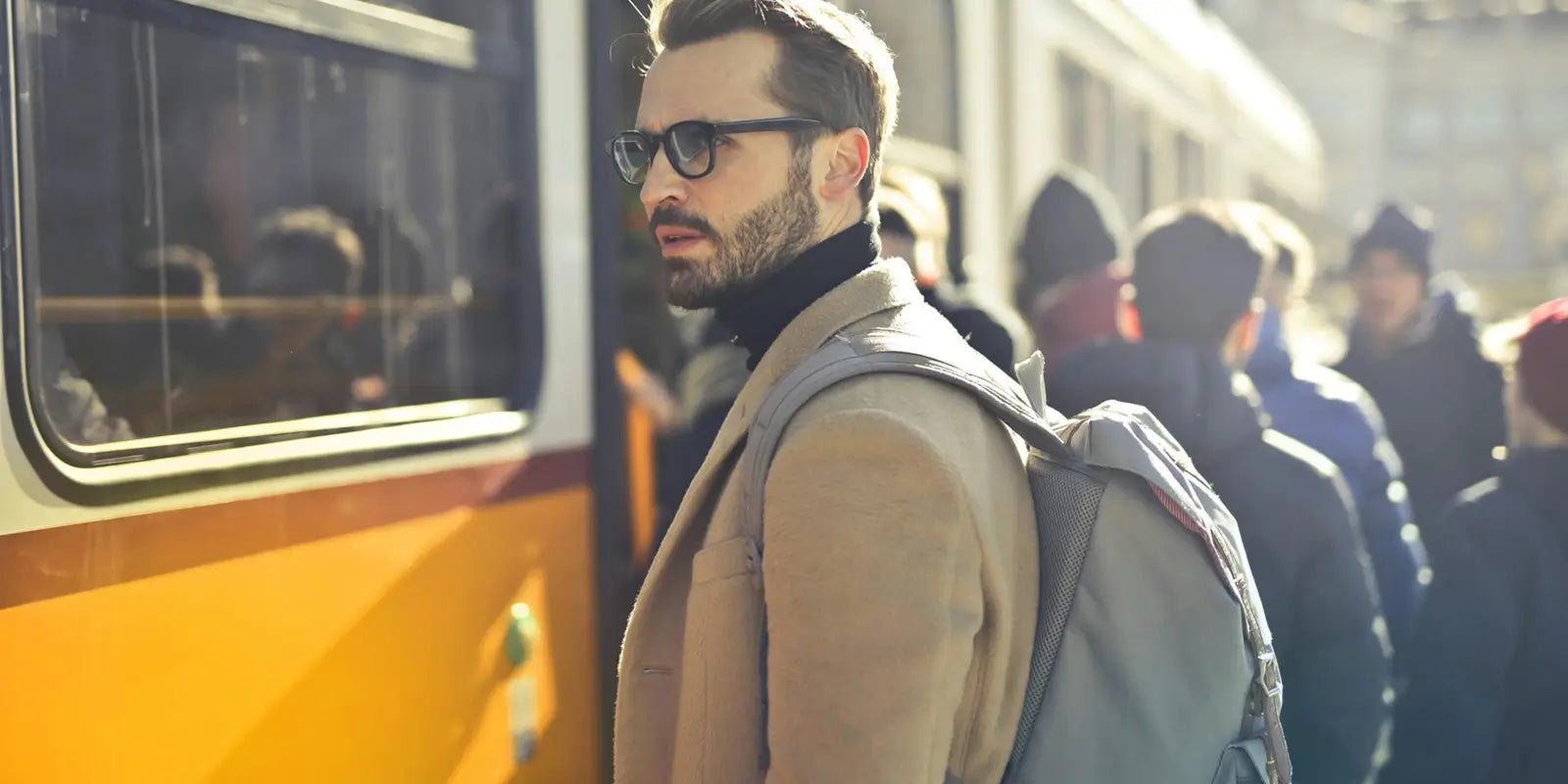 Homme barbu élégant portant un manteau beige et un sac à dos homme gris, attendant devant un tramway dans une rue animée, illustrant un quotidien urbain dynamique et connecté.