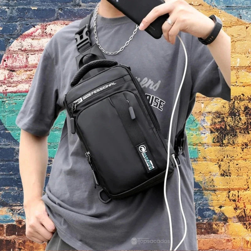 Petit sac à dos homme noir porté en bandoulière par un homme en t-shirt gris, en train de charger son téléphone en extérieur devant un mur coloré.