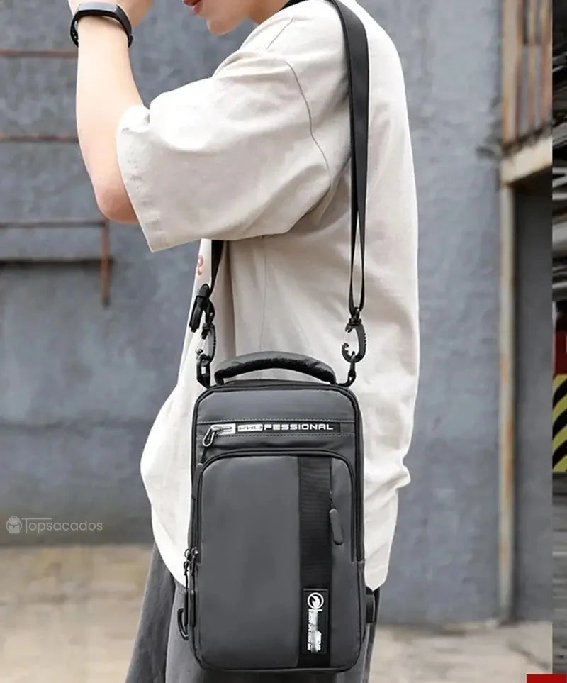 Petit sac à dos homme porté en bandoulière par un homme en t-shirt clair dans une ruelle extérieure, avec mousquetons visibles sur les sangles.