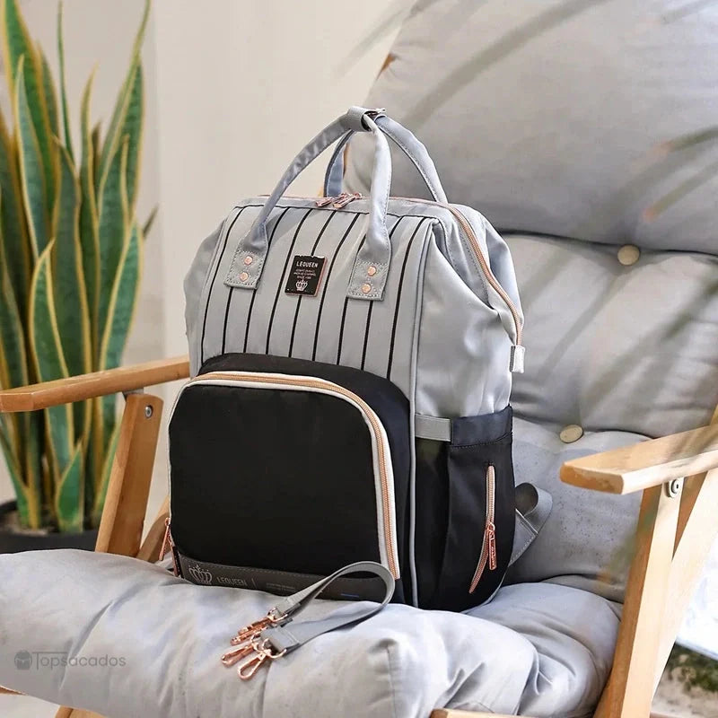 Sac à dos à langer Urbain et chic posé sur un fauteuil en bois et coussin gris, dans un intérieur lumineux avec plante décorative.