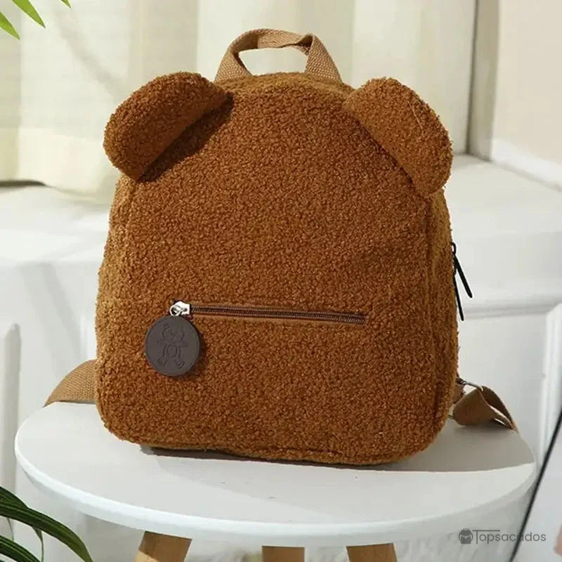 Sac a dos college Ourson marron effet peluche avec oreilles rondes, présenté sur tabouret blanc dans une pièce cosy