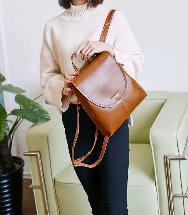 Sac à dos cuir femme marron porté à la main par une femme en pull crème dans un salon lumineux.