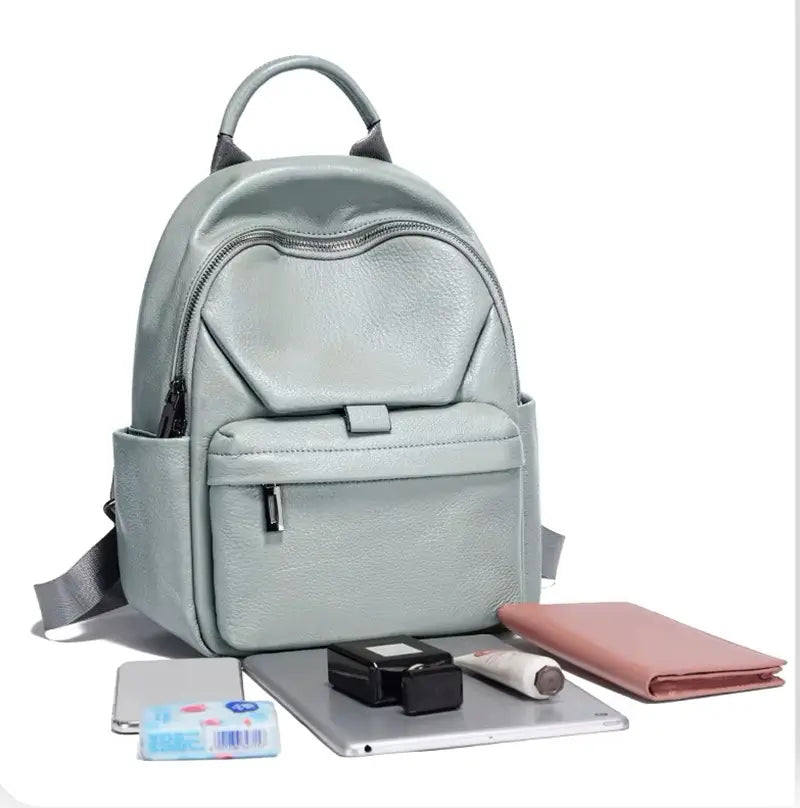 Sac à dos cuir femme vert clair avec ses accessoires du quotidien posés devant : tablette, portefeuille rose, baume, étui noir et paquet de mouchoirs.