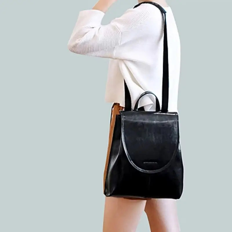 Sac à dos femme en cuir noir porté à l’épaule par une femme en jupe beige et pull blanc sur fond uni, soulignant son élégance moderne.