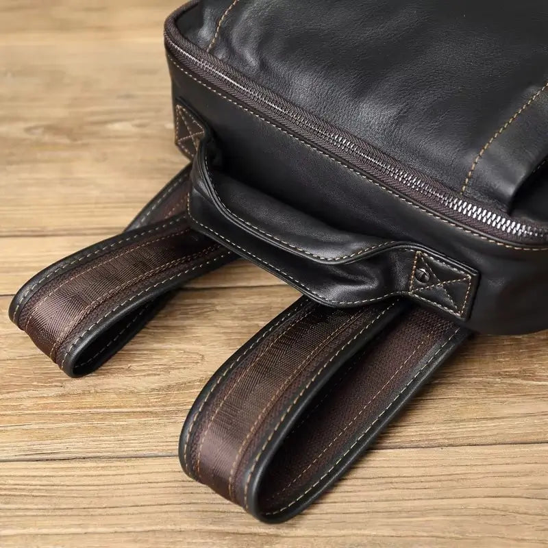 Sac à dos homme cuir noir détail des sangles d'épaule épaisses et rembourrées, posées sur plancher en bois clair.